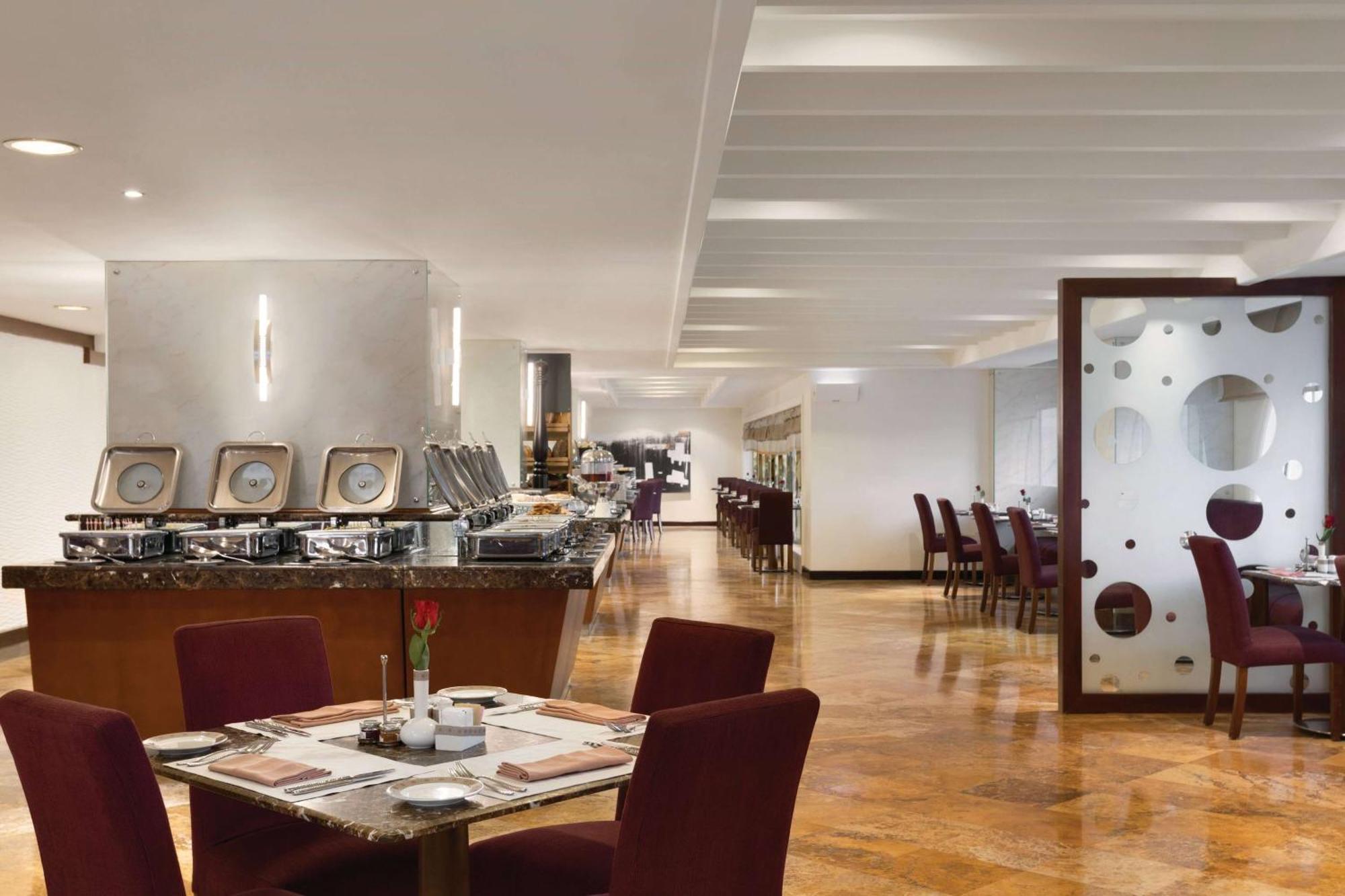 Grand Regency Doha, Trademark Collection By Wyndham Exterior foto The photo shows a modern restaurant interior. There are several tables with maroon chairs arranged throughout the space. One table is set with a small vase containing a flower and dining utensils. Along one side, there is a buffet area with a sleek c