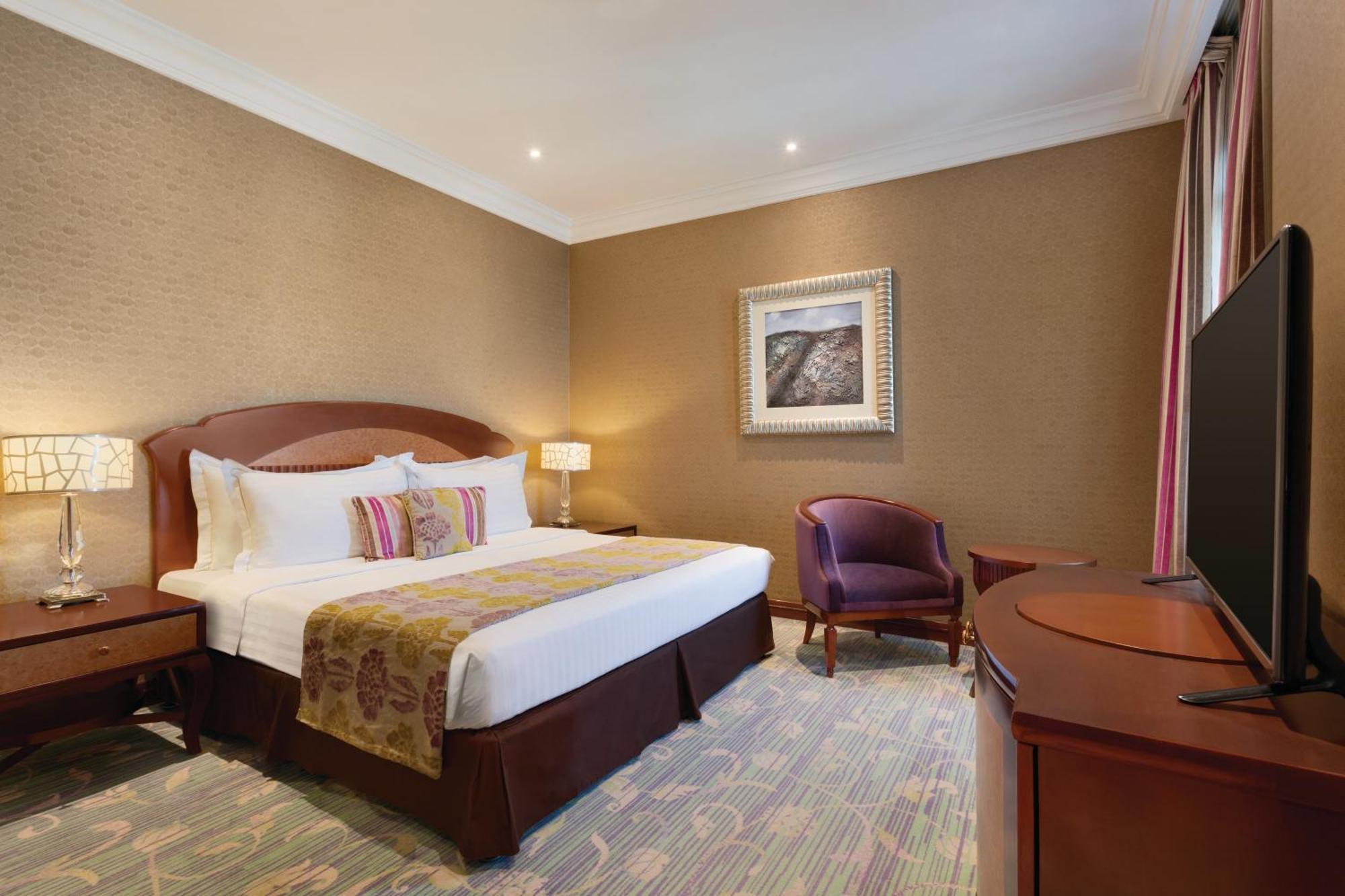 Grand Regency Doha, Trademark Collection By Wyndham Exterior foto The image shows a hotel room that features a cozy and elegant design. There's a large bed with white sheets and colorful decorative pillows. Next to the bed, there are nightstands with lamps. A comfortable armchair is positioned in the corner, and a 