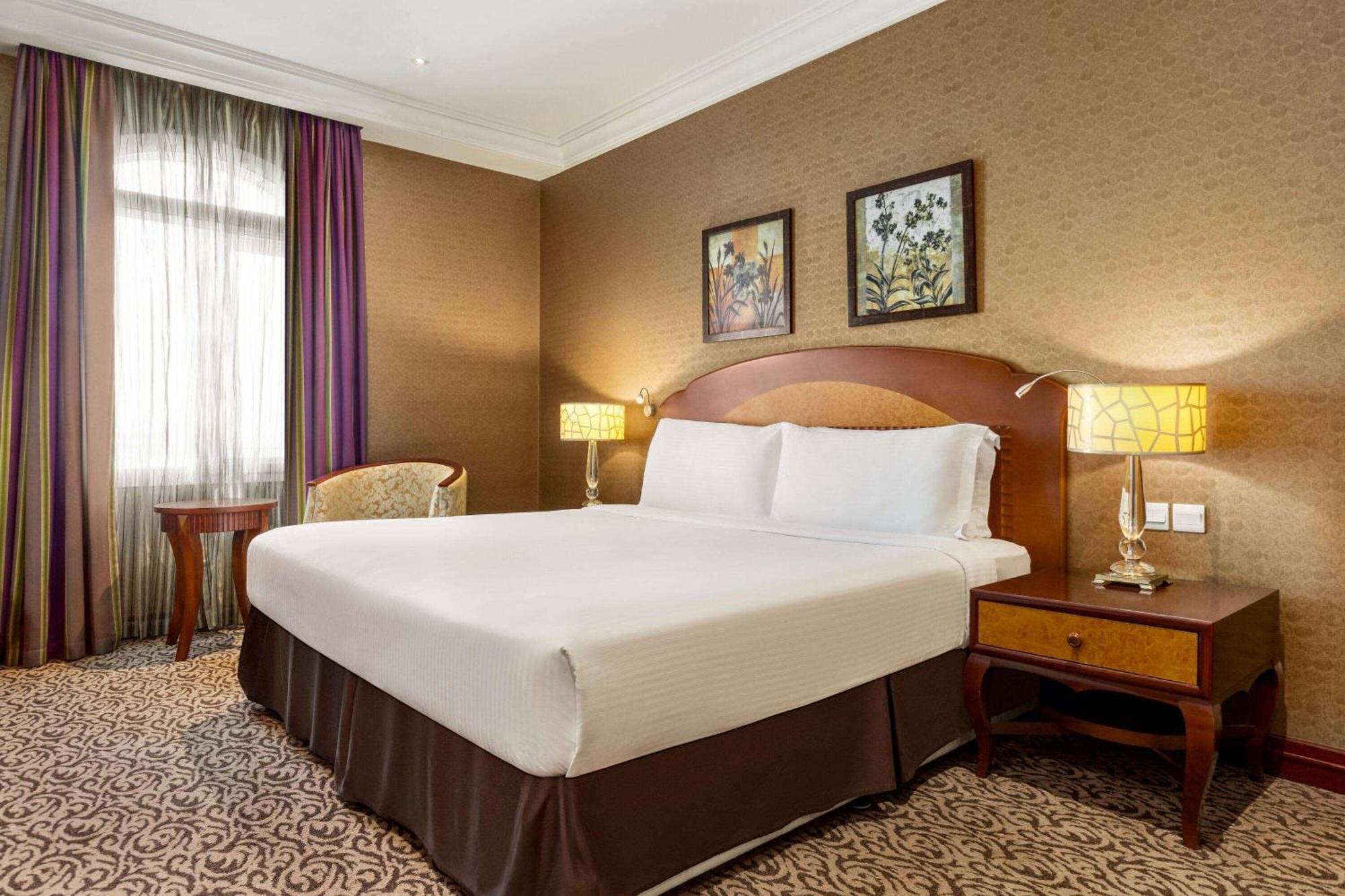 Grand Regency Doha, Trademark Collection By Wyndham Exterior foto The photo displays a comfortable hotel room featuring a large bed with white linens and a brown bed skirt. The room has a warm color palette, with textured brown walls and patterned carpet. There are two bedside lamps providing soft lighting next to 